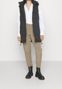 Svart pufferväst med huva, med en quiltad design och blixtlås, bärs över en vit skjorta och beiga byxor, i kombination med svarta stövlar.