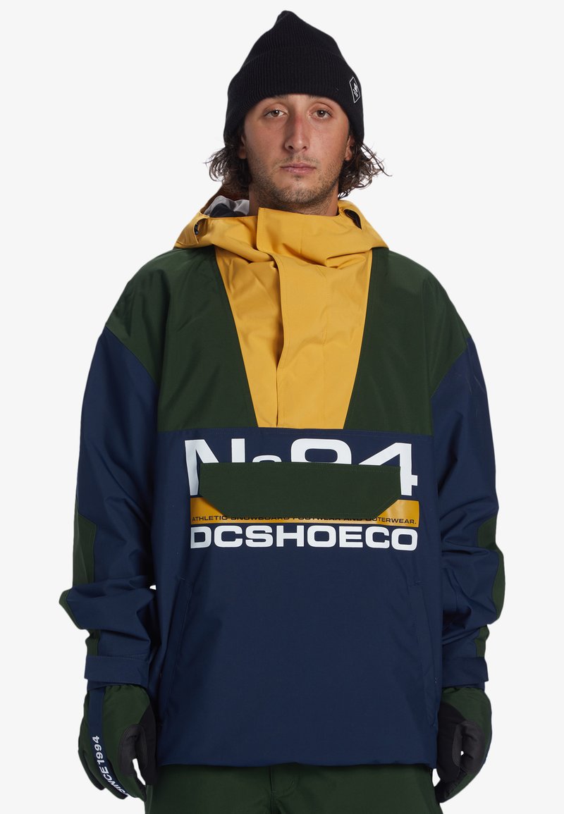 Color-blocked jacket in navy, green, and yellow. Features bold logos, a kangaroo pocket, and a high collar with an adjustable hood.