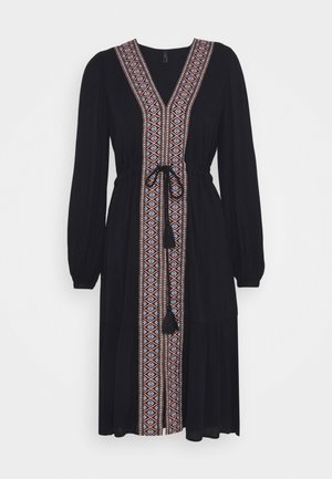 Černé šaty s dlouhými rukávy a hlubokým výstřihem do V, se barevným geometrickým vzorem na přední části a páskem na zavázání v oblasti pasu.