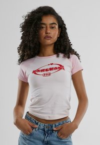 Fitted tee with short pink sleeves and white body, featuring a red logo and heart accents. Paired with high-waisted light blue jeans.