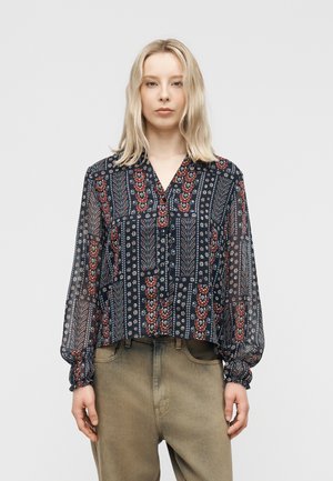 Lyshåret kvinde iført en marineblå gennemsigtig bluse med røde og hvide blomsterprint og beige bukser med høj talje mod en ensfarvet baggrund.