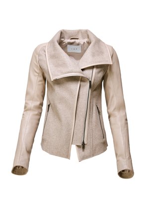 Femme portant une veste beige, un pantalon gris asymétrique, tenant un sac à main noir avec un accessoire en fourrure, debout devant un fond clair uni.
