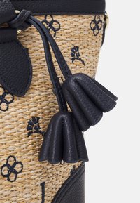 Sac en paille tressée avec des accents en cuir bleu marine, ornés de pompons en cuir décoratifs et de broderies florales en bleu marine sur fond beige.