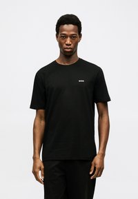 Νεαρός άνδρας στέκεται μπροστά σε απλό ανοιχτό φόντο, φοράει μαύρο t-shirt BOSS και μαύρο παντελόνι, κοιτάζοντας μπροστά με ουδέτερη έκφραση.