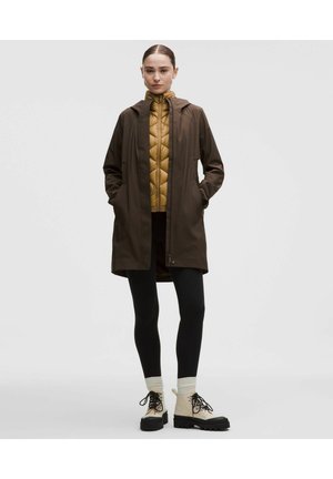 Frau trägt einen braunen Kapuzenmantel über einer senffarbenen gesteppte Jacke, schwarze Leggings, cremefarbene Socken und beige Schnürstiefel, steht mit den Händen in den Taschen.