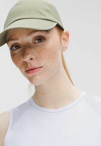 Jonge vrouw met sproeten die een lichtgroen petje en een witte mouwloze top draagt, recht vooruitkijkend met een neutrale uitdrukking.