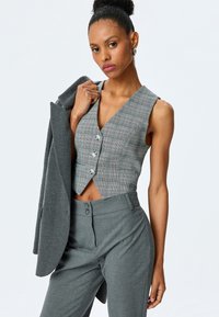 Grau karierter taillierter Weste mit drei Knöpfen, kombiniert mit grauen, maßgeschneiderten Hosen. Ein passender grauer Blazer liegt über dem Arm. Glatte Stofftextur.