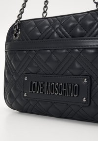 Melna kvadrāta ādas rokassoma ar ķēdes siksnu, kurai ir metāla plāksne ar uzrakstu "LOVE MOSCHINO" un dekoratīviem kniedēm.