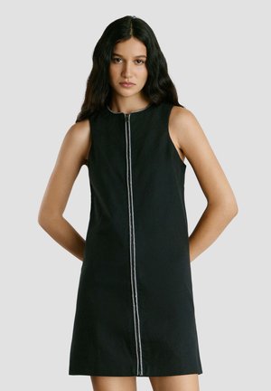 Vrouw met lang donker haar, gekleed in een mouwloze zwarte jurk met ritssluiting aan de voorkant, staand met handen achter de rug tegen een lichtgrijze achtergrond.