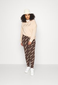 Beige džemperis ar "adidas" logo, brūni zīmējuma legingi ar atkārtojošos zīmola dizainu, balti kedu un krēmkrāsas buckets cepure.