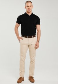 Férfi áll, jobb kezét a zsebében tartja, fekete galléros pólót, bézs nadrágot, barna övet, barna cipőt és karórát visel, világos háttér előtt.