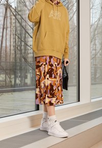 Khaki hoodie with front pocket and logo, paired with a patterned knee-length skirt featuring purple, brown, and cream colors. White sneakers complete the look.