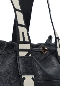 Borsa in pelle nera con superficie testurizzata, dotata di una larga tracolla crema con lettering nero e un'accento in hardware dorato all'apertura.