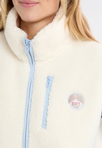 Kurtka polarowa w kremowym kolorze z wysokim kołnierzem, z jasnoniebieskimi zamkami błyskawicznymi i naszywką z wzorem góry i gwiazdy na piersi.