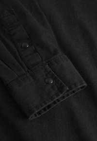 Polsino di camicia in tessuto nero con una superficie liscia, caratterizzato da un design a punta e due bottoni a pressione neri. Primo piano del bordo del polsino arrotolato.