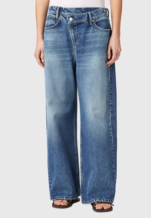 Person iført løstsiddende, højtaljede blå jeans med falmede områder, kombineret med sorte sandaler med åbne tæer mod en ensfarvet baggrund.