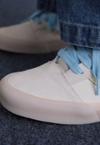 Bijele niske tenisice od suede i mrežastog materijala, svijetloplave vezice, okrugli vrh i suptilni detalji šavova na tamnoj tepih pozadini.