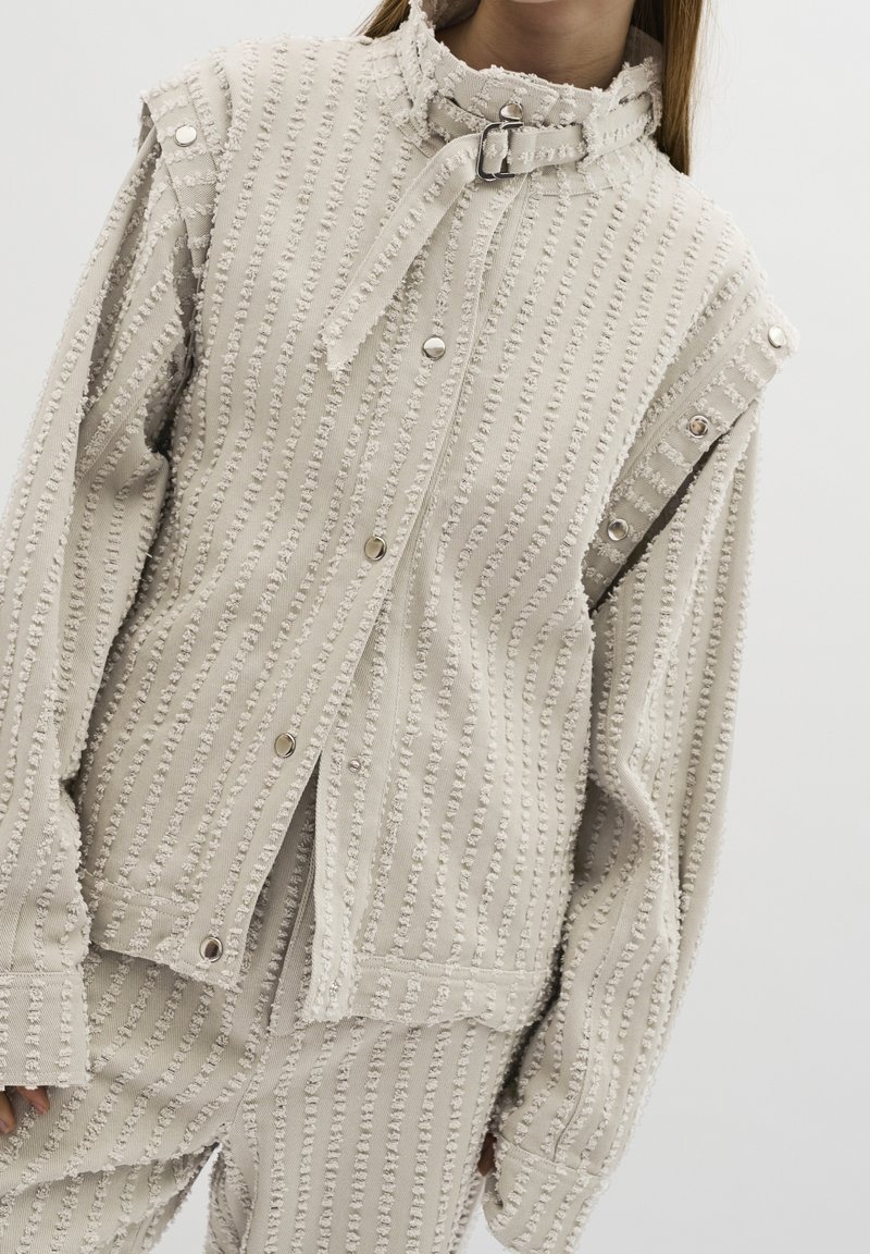 Harmaa tekstiili takki kohotetulla raidalla, painonappikiinnitys, solkakaulus, ja siihen sopivat housut päällä henkilö, yksinkertaisen valkoisen taustan edessä.