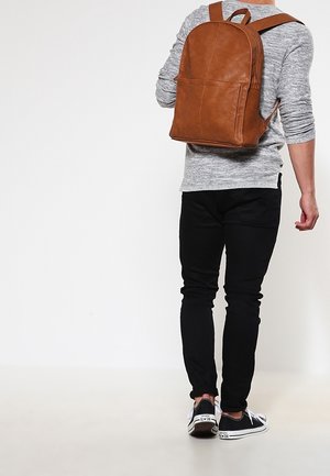 Personne portant un pantalon noir, un pull gris, des baskets noires, avec un sac à dos en cuir marron, debout sur un fond blanc uni.