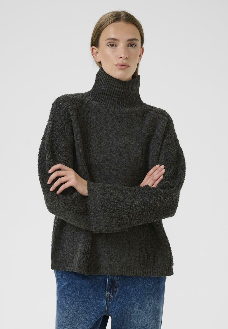 Una mujer de piel clara y cabello castaño viste un suéter de cuello alto texturizado en gris oscuro y jeans azules, de pie con los brazos cruzados.
