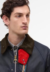 Ναυτικό σακάκι με κολάρο από καστόρι, κόκκινη υφασμάτινη λεπτομέρεια και φερμουάρ με ριγωτή λαβή. Φοράει από κάτω ένα καρό πουκάμισο.