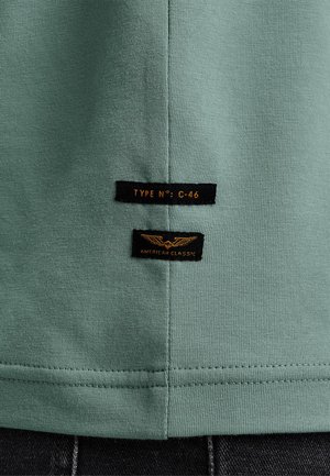 Zeleni bombažni majica z gladko teksturo, ki ima dve črni nalepki z napisi "TIP ŠT: C-46" in "AMERIŠKA KLASIČNA" v oranžni pisavi.