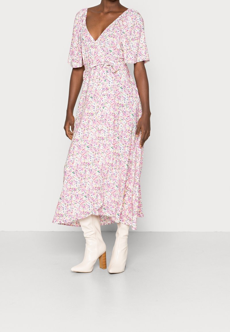 Femme portant une robe midi fleurie rose à manches courtes et des bottes blanches à talons hauts, debout sur un sol gris.