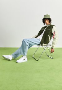 Giacca varsity verde e bianca, jeans azzurri chiari, sneakers bianche con dettagli neri e un cappello a secchiello verde, seduto su una sedia pieghevole in cromo.