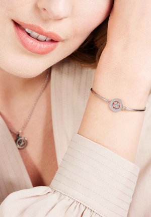 Zilveren armband met een ronde hanger met een uitsnede in bloemmotief. Draagt een lichte, gestructureerde blouse.