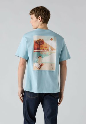 Young man wearing light blue T-shirt with a colorful poolside lounge graphic on the back, paired with dark blue jeans.