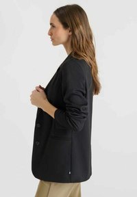 Femme aux longs cheveux bruns, portant un blazer noir et un pantalon beige, se tenant de profil devant un fond clair et uni.