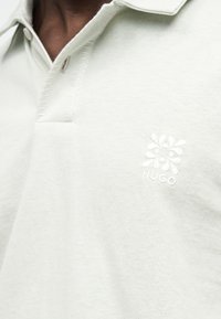 Κοντινό πλάνο ενός ατόμου που φοράει μια κρεμ πόλο με λουλουδάτο λογότυπο και τη λέξη «HUGO» κεντημένη στο στήθος.