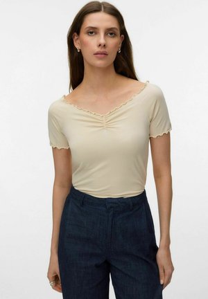 Donna che indossa una maglia beige a maniche corte arricciate con rifinitura a bordo ondulato e pantaloni a vita alta blu scuro, in piedi davanti a uno sfondo semplice.