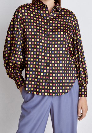 Vrouw draagt een blouse met lange mouwen en knopen, met een patroon van gele, rode en blauwe vierkanten, gecombineerd met lichtpaarse broek.