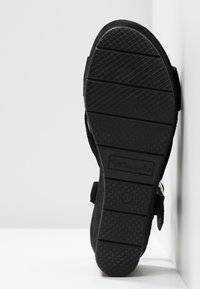 Semelle noire de sandale avec grip texturé et nom de la marque "Tamaris," affichée à la verticale sur un fond blanc de mur et de sol.