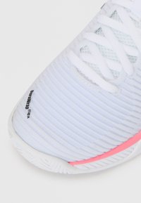 Hvit sportsko med en teksturert overflate, som har horisontale striper, rosa detaljer på sålen, og hvite lisser. Holdbart materiale vist.