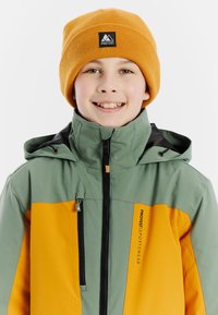 Groen en oranje bicolor jack met hoge kraag, voorrits en logo. Draagt een mosterdkleurige beanie, wat een sportief ontwerp laat zien.