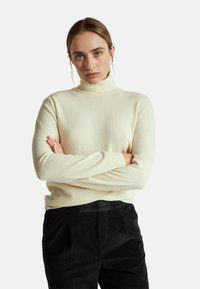 Maglione a collo alto color crema realizzato in morbido materiale, con maniche lunghe e design aderente, indossato con pantaloni in velluto a coste neri.