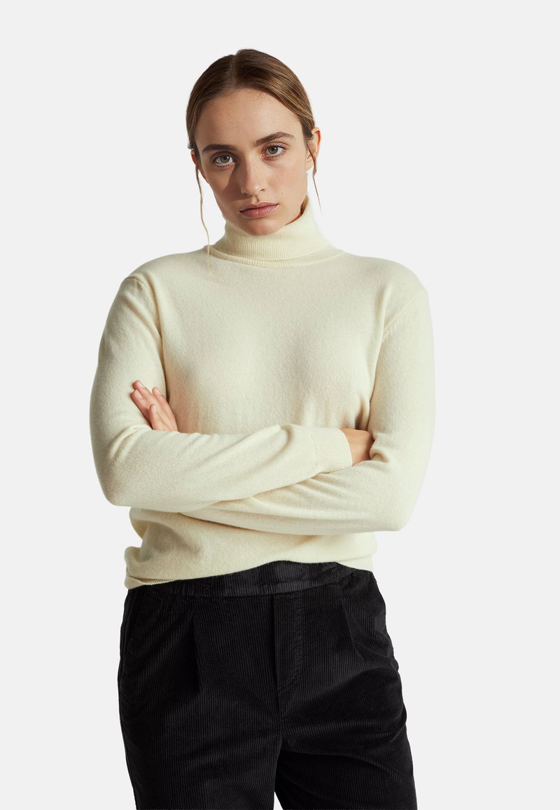 Maglione a collo alto color crema realizzato in morbido materiale, con maniche lunghe e design aderente, indossato con pantaloni in velluto a coste neri.