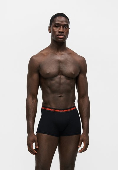 Homme musclé portant un boxer noir avec une ceinture rouge, debout devant un fond clair uni, faisant face.
