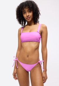 Γυναίκα με σγουρά μαλλιά, που φοράει φωτεινό ροζ μπικίνι με πλευρικούς ρυθμιζόμενους δεσίμους και φαρδιές τιράντες στους ώμους, που στέκεται μπροστά σε απλό φόντο.
