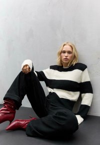 Pull à rayures noir et blanc avec une coupe décontractée, associé à un pantalon noir ample et des bottes rouges à bout pointu. Texture lisse, design décontracté.
