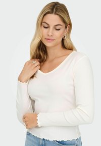 Blondynka z falującymi włosami, ubrana w białą, prążkowaną bluzkę z długim rękawem i falbaniastymi brzegami oraz jasnoniebieskie dżinsy, delikatnie patrząca w dół.