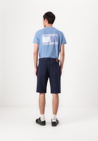 Lyseblå bomull T-skjorte med et trykt firkantet design på baksiden, kombinert med marineblå shorts og svarte joggesko.