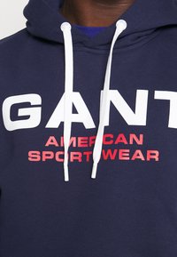 Sudadera con capucha azul marino con cordones blancos. Presenta un texto audaz en blanco y rojo: "GANT AMERICAN SPORTWEAR." Material de algodón suave y diseño relajado.
