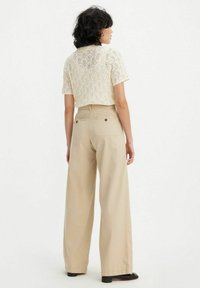 Strickcreme-T-Shirt mit kurzem Arm und strukturiertem Muster, kombiniert mit hoch sitzenden, weit geschnittenen beigefarbenen Hosen, die über rückseitige Taschen und Gürtelschlaufen verfügen.