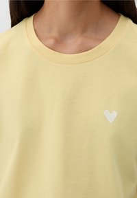 Camiseta de algodón amarilla con un clásico escote redondo y mangas cortas. Presenta un pequeño corazón bordado en blanco en el lado izquierdo del pecho.