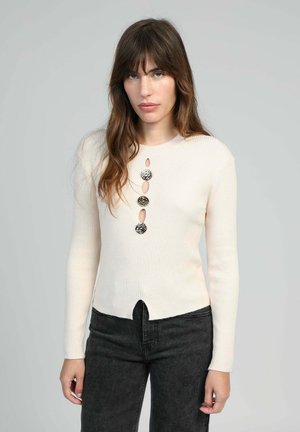Femme avec de longs cheveux bruns portant un pull crème à côtes avec trois grands boutons ronds noir et blanc et un jean noir contre un fond uni.