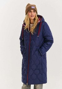 Granatowy pikowany długi płaszcz z kapturem, bordowym zamkiem błyskawicznym i sznurkiem do ściągania. Posiada boczne kieszenie oraz podszewkę w kwiatowy wzór.