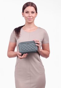 Prošívaná zelená kožená peněženka se zipovým uzávěrem, obdélníkového tvaru, s jemným logem a texturovaným vzorem na povrchu.